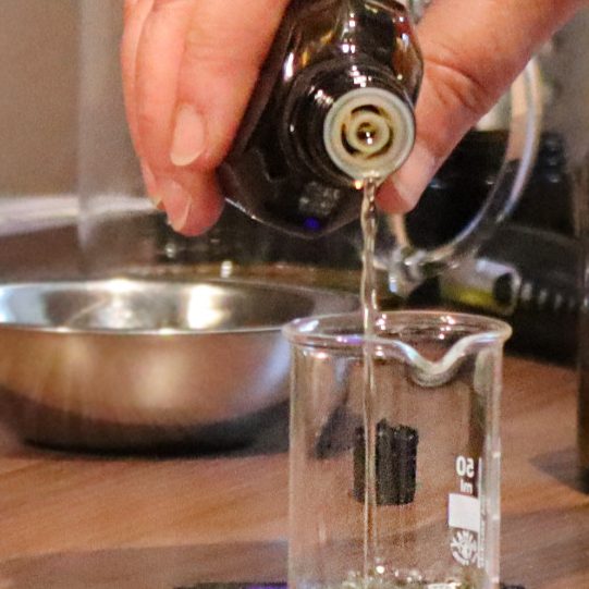 Natürliche Zutaten Eine Hand hält eine braune Glasflasche aus der eine klare Flüssigkeit in ein durchsichtiges Glas rinnt.