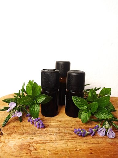 Drei schwarze Fläschchen mit Kräutern und lila Blüten auf einem Holztisch.