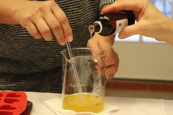Hände geben Flüssigkeit aus einer Flasche in einen Messbecher mit gelber Flüssigkeit.