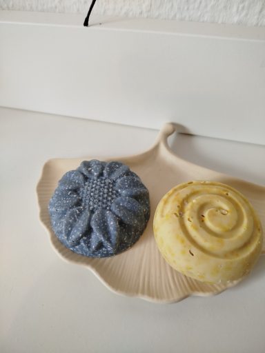 Zwei Seifen: eine blaue in Sonnenblumenform und eine runde gelbe mit Spiralmuster auf einem weißen Teller in Form eines Gingkoblattes