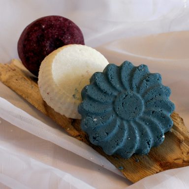 Drei farbige, runde Shampoobars auf einem Holzbrett: blau, weiß und rot.