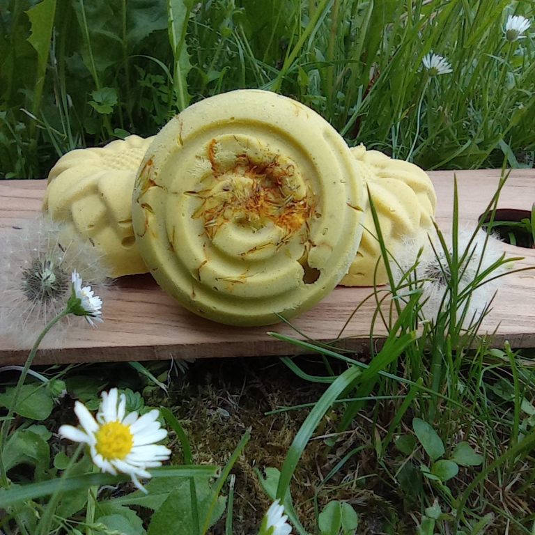 Ringelblumenseife Runde hellgelbe Seife mit Spiralförmiger Prägung auf einem Holzbrett, im Hintergrund zwei weitere Seifen und Wiese.
